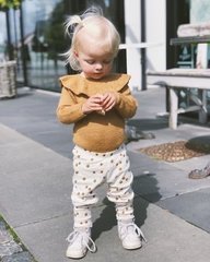 Lille pige med lyst hår i hestehale iført brun bluse med flæsekrave og hvide bukser med sorte pletter, stående udendørs på en flisebelagt terrasse. (2)