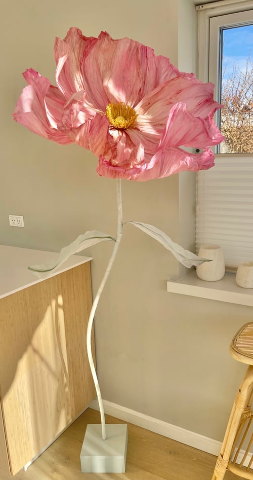 Stor, lyserød kunstig blomst med gult centrum på lang hvid stilk med blade, placeret i et lyst rum ved et vindue. (1)
