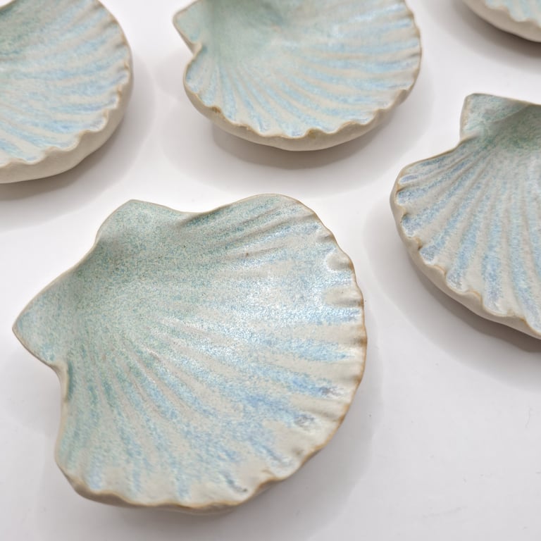 Små keramiske skåle formet som muslingeskaller med lyseblå og cremefarvede nuancer, placeret på en hvid overflade.