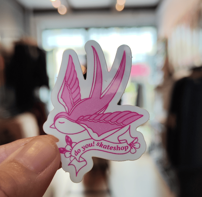 Sticker med en pink fugl og teksten "do you! skateshop" holdt mellem to fingre i en butik.
