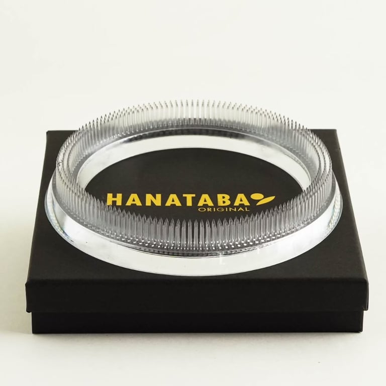 Klar, cirkulær Hanataba blomsterholder med spidse nåle, placeret på en sort æske med gul tekst.