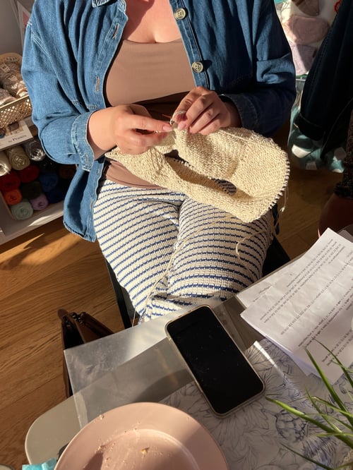 En person iført en blå denimjakke og stribede bukser strikker med beige garn, mens de sidder på en stol. Et bord foran dem har en smartphone, papirer og en tom lyserød tallerken. I baggrunden ses en hylde med forskellige farver garn. (4)