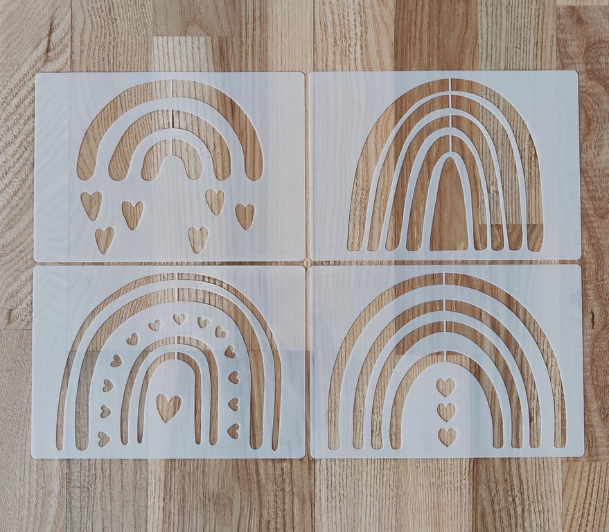 Fire hvide malerstencils med forskellige regnbuedesigns og hjerter skåret ud, lagt på et lyst træbord.