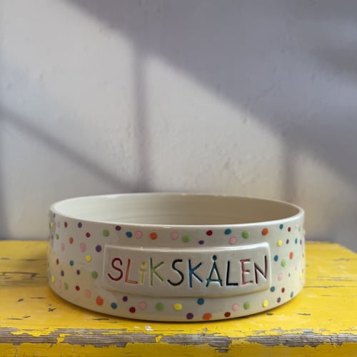 En hvid skål med farverige prikker og teksten "Slikskålen" i forskellige farver, placeret på et slidt, gult bord mod en grå væg. (1)
