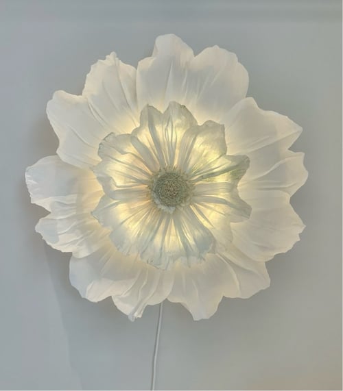 Hvid blomstervæglampe med bløde, gennemsigtige kronblade, der lyser indefra mod en lys væg. (1)