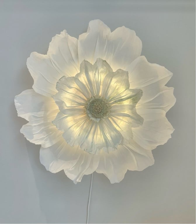 Hvid blomstervæglampe med bløde, gennemsigtige kronblade, der lyser indefra mod en lys væg.
