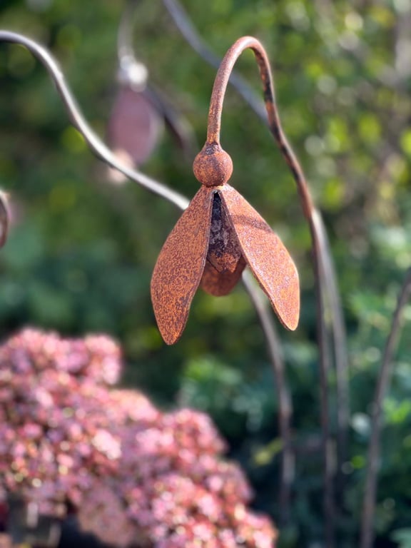 Rustent metalornament formet som en hængende blomst med bladlignende dele i nærbillede, med grønne og rosa slørede planter i baggrunden.