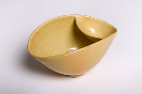 En beige, keramisk skål med en lille, opadgående tud, placeret på en hvid baggrund.