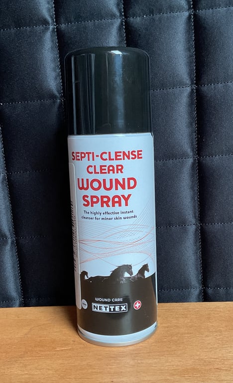 Flaske med Septi-Cleanse Clear Wound Spray, et desinfektionsspray til mindre hudsår, stående på en træoverflade med sort quiltet baggrund.