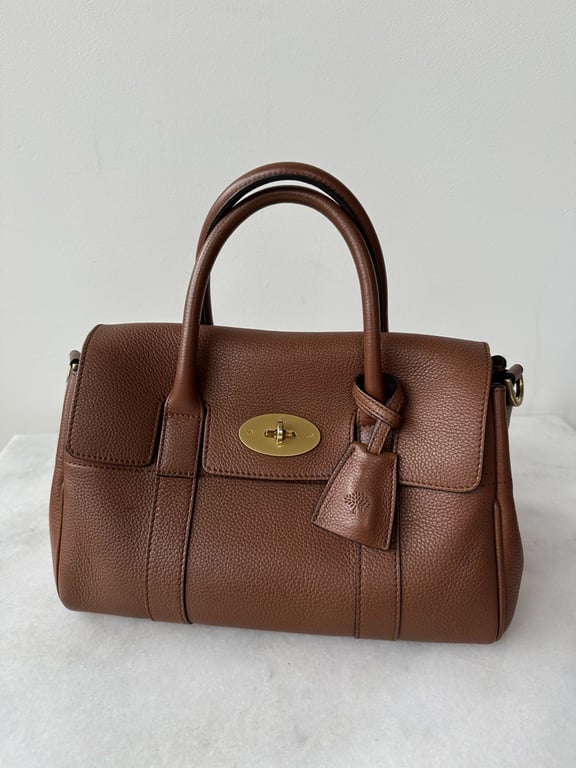 Brun lædertaske med to håndtag og guldfarvet spænde, placeret på hvid overflade foran hvid væg.