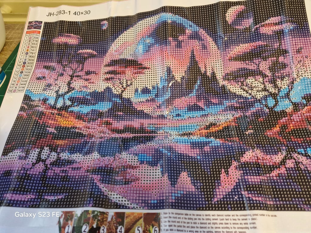Diamond painting med et farverigt naturscene-motiv, der viser et bjerglandskab under en stor måne og lyserøde blomstertræer, med symboler og farvekoder på lærredet.
