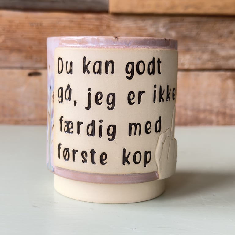 Keramikkrus med tekst på dansk: "Du kan godt gå, jeg er ikke færdig med første kop".
