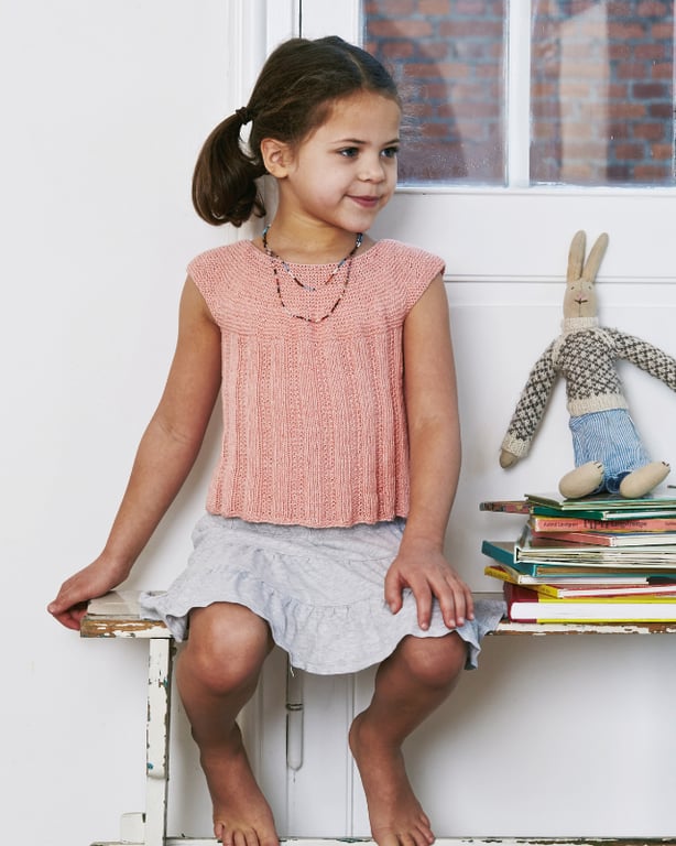 En lille pige med mørkt hår, iført en lyserød strikket top og en lysegrå nederdel, sidder barfodet på en bænk ved siden af en stak bøger og en blød bamse i form af en kanin.