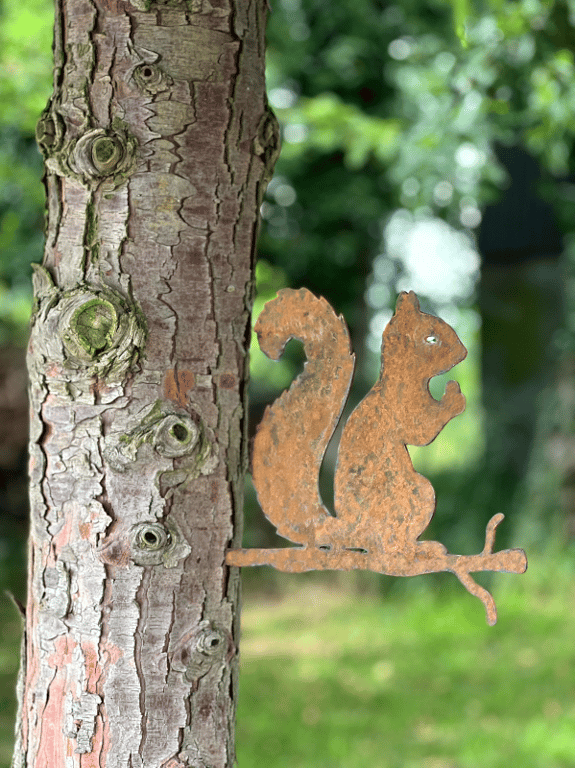 Rustent metalornament formet som et egern, der sidder på en gren, ved siden af en træstamme med knuder i barken.