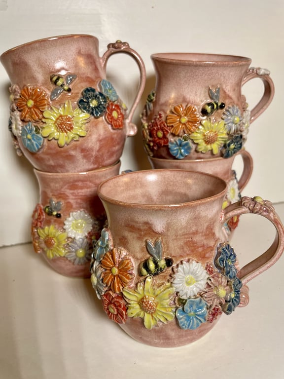 Billede af fire rosefarvede keramikkrus med relief af farverige blomster og bier rundt om koppen.