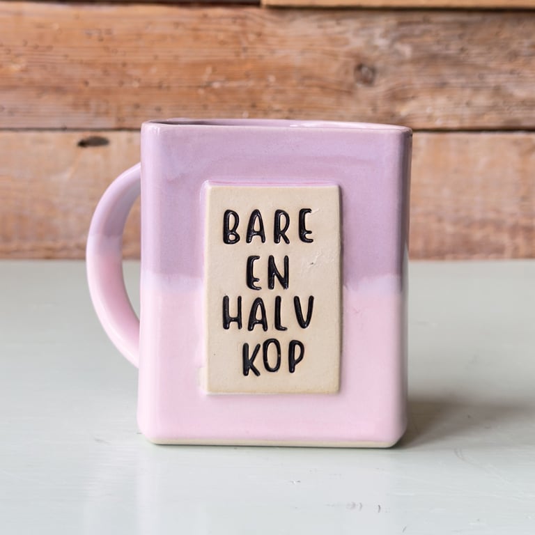 Lyserød keramisk kop med tekst "Bare en halv kop" på forsiden, placeret på en lys overflade med en træbaggrund.