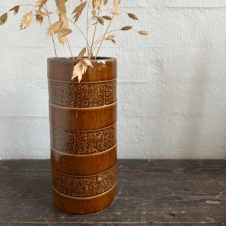 Brun keramisk vase med tørrede græsstrå stående på et mørkt træbord foran en lys væg.