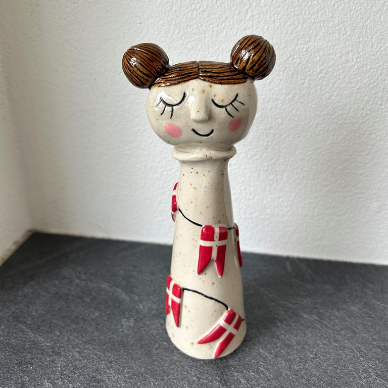Keramisk figur af en pige med to knolde i håret, lukkede øjne, røde kinder og en krave, dekoreret med danske flag på en sort baggrund.