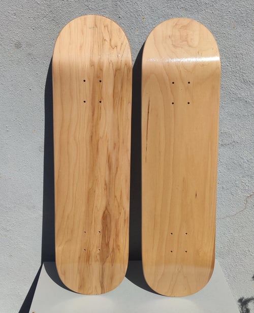 To tomme skateboard decks i lyst, naturligt træ, placeret lodret mod en grå betonvæg. (1)
