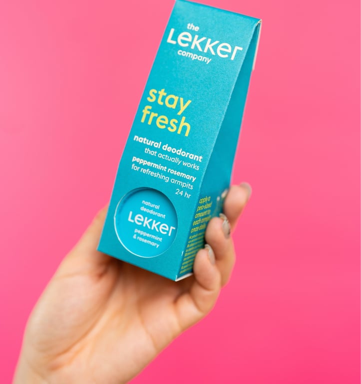 Hånd, der holder en blå æske med naturlig deodorant fra The Lekker Company med pebermynte og rosmarin mod en pink baggrund.
