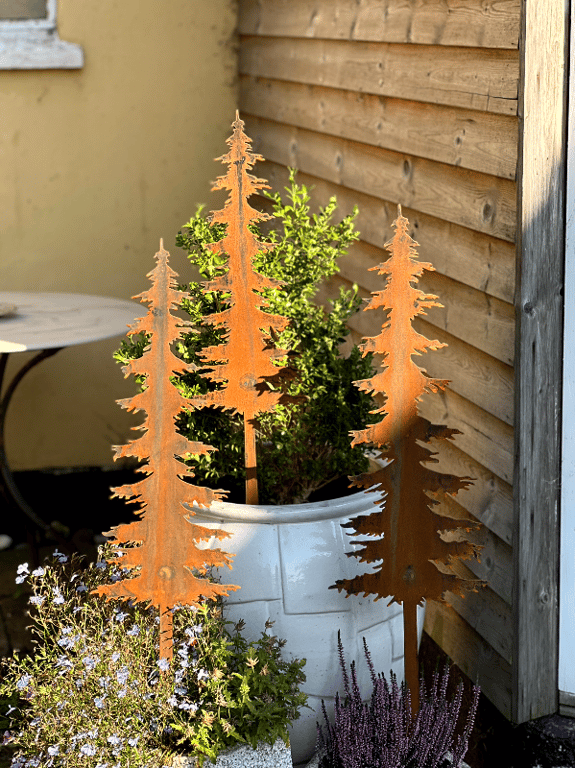 Tre rustne metaldekorationer formet som grantræer placeret i en hvid urtepotte med planter, op ad et træbeklædt væg.