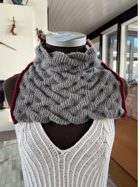 Mannekeng iført hvid, ribstrikket top og en grå, håndstrikket halsvarmer med mønster og rød kant.