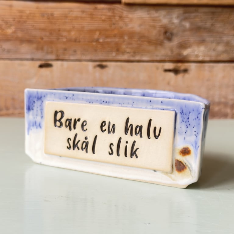 Keramisk skilte med blå kanter og tekst på dansk: "Bare en halv skål slik", placeret på hvid bordplade med træbaggrund.