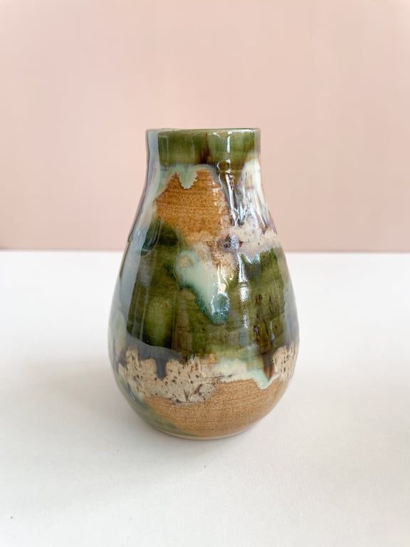 Keramisk vase med grønne, beige og hvide glasurdetaljer, placeret på en hvid overflade med lyserød baggrund.