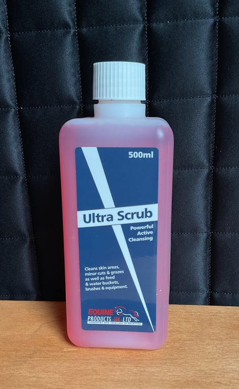 Flaske med 500 ml Ultra Scrub rengøringsmiddel til hud, snit og udstyr, stående på træbord foran sort quiltet baggrund.