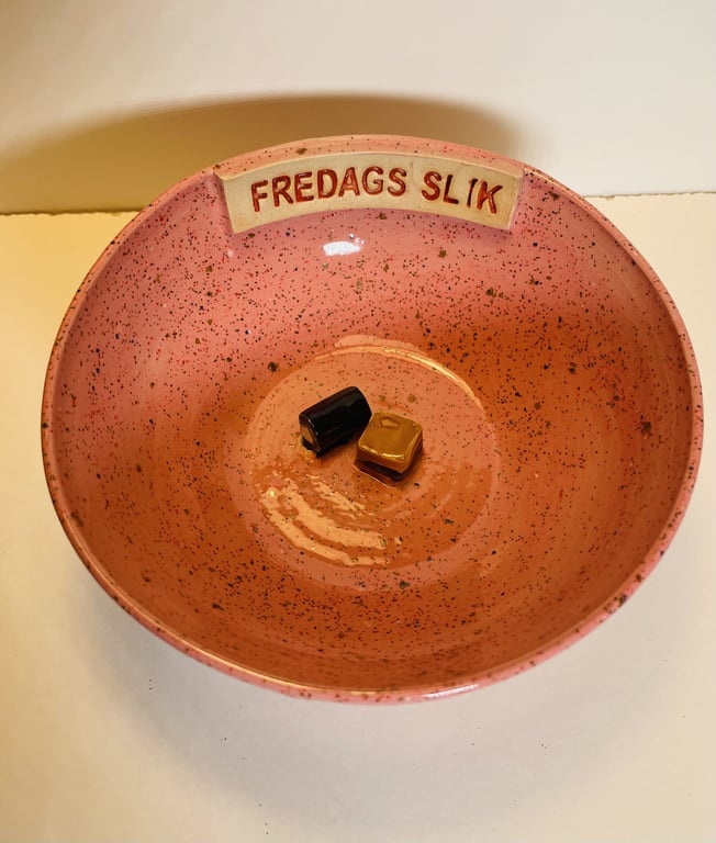 Lyserød skål med prikker og teksten "FREDAGS SLIK" med to små karameller indeni.