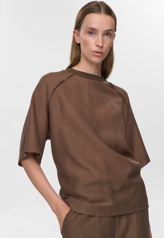 Kvinde iført løs brun bluse med korte ærmer og matchende bukser, stående med hånden i lommen mod hvid baggrund.
