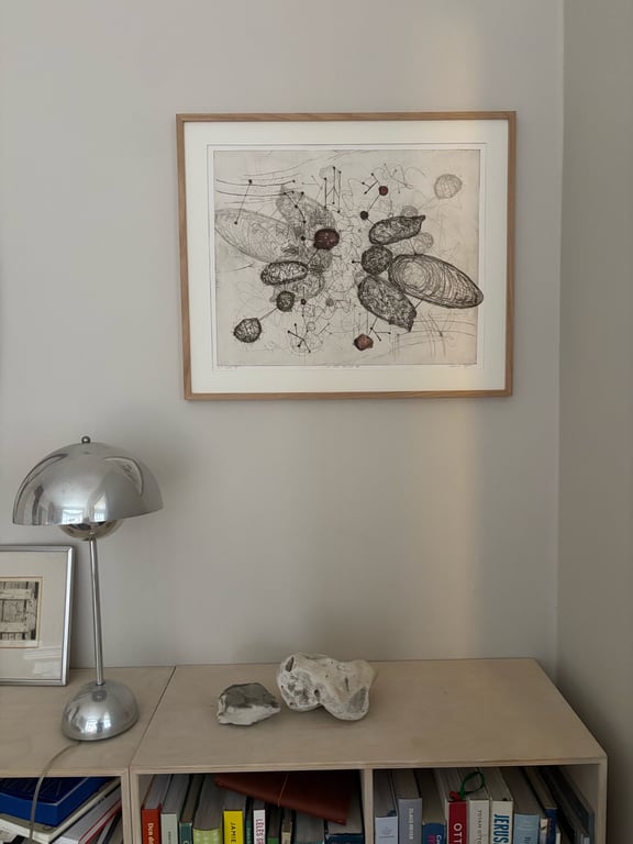 Tegning med abstrakte, organiske former i sort og brun indrammet og hængt på hvid væg over et lyst møbel med to sten, en sølvfarvet bordlampe og bøger.