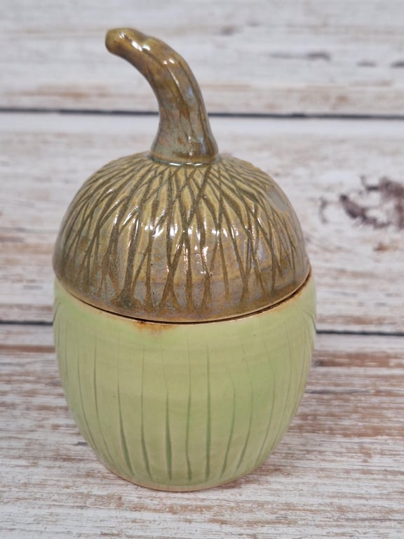 Keramisk beholder formet som et agern med brun låg og grøn bund, placeret på trægulv.