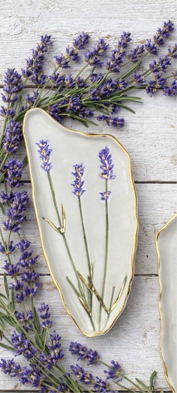 Lille hvid keramisk fad med guldkanter og malede lavendelblomster, omgivet af friske lavendelgrene på hvid træoverflade.