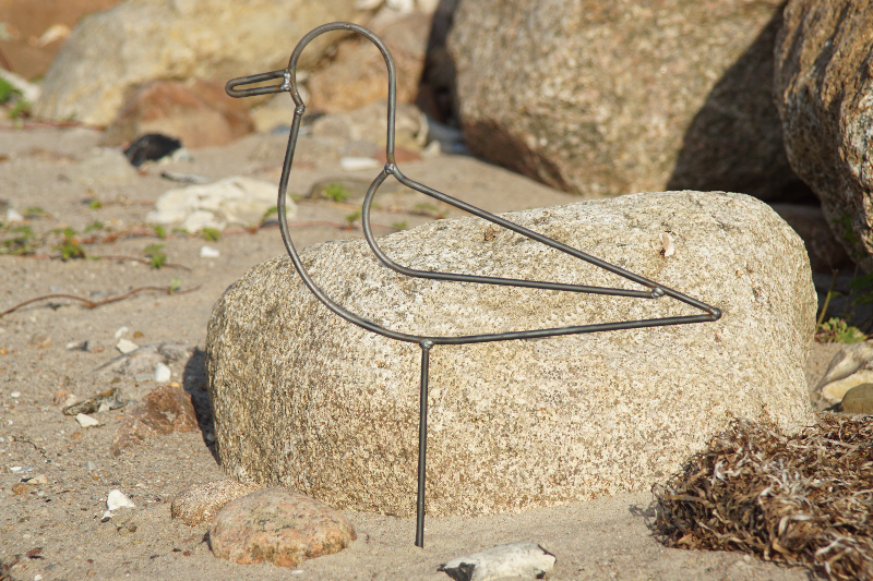 Metallinspireret fugleskulptur lavet af sort tråd, placeret på en sandstrand med store sten i baggrunden.