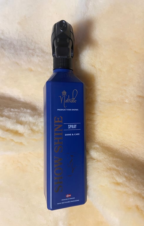 Flaske med blå spraydåse mærket "Nathalie Show Shine Spray" til glans og pleje, placeret på en beige blød overflade.