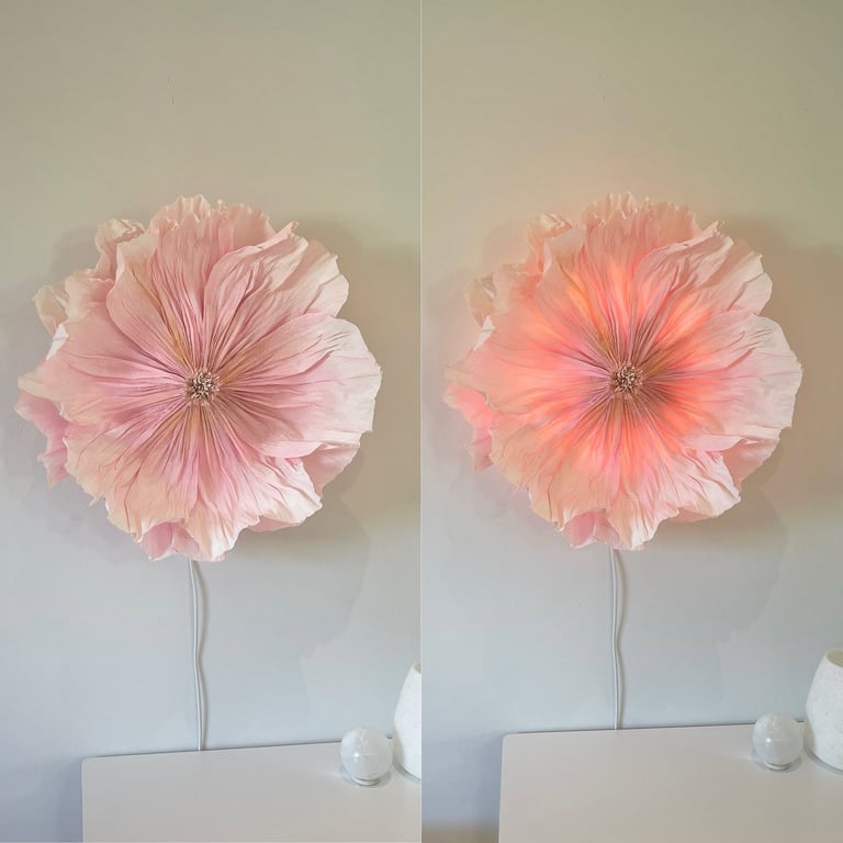 To billeder ved siden af hinanden af en stor, lyserød blomsterlampe monteret på en væg over et hvidt bord. Til venstre er lampen slukket, til højre er den tændt og udsender et varmt, orange lys indvendigt.