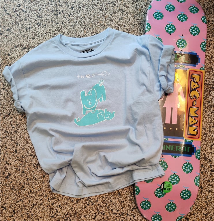 Lyseblå T-shirt med tegneserieprint af to dyr og teksten "there" pålagt en lyserød skateboard med grønne prikker og Pac-Man-logo, der ligger på en marmoreret gulvflise.