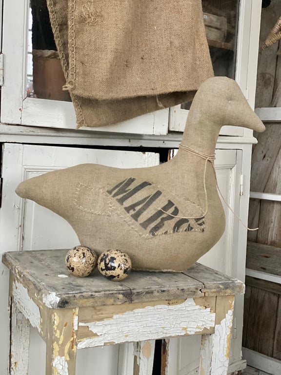 En dekoration af en gås lavet af groft stof med teksten "MARC" på siden, placeret på et gammelt, afskallet træbord med to plettede æg foran, i et rustikt rum med hvidmalede møbler og hængende jutesæk.