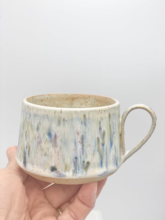 Hånd, der holder en keramisk kop med glasur i hvide, blå, grønne og røde nuancer på en lys baggrund.