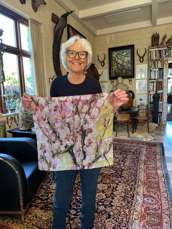 Ældre kvinde med kort gråt hår og briller står i et hjemligt rum og holder et tørklæde med blomsterprint frem mod kameraet.