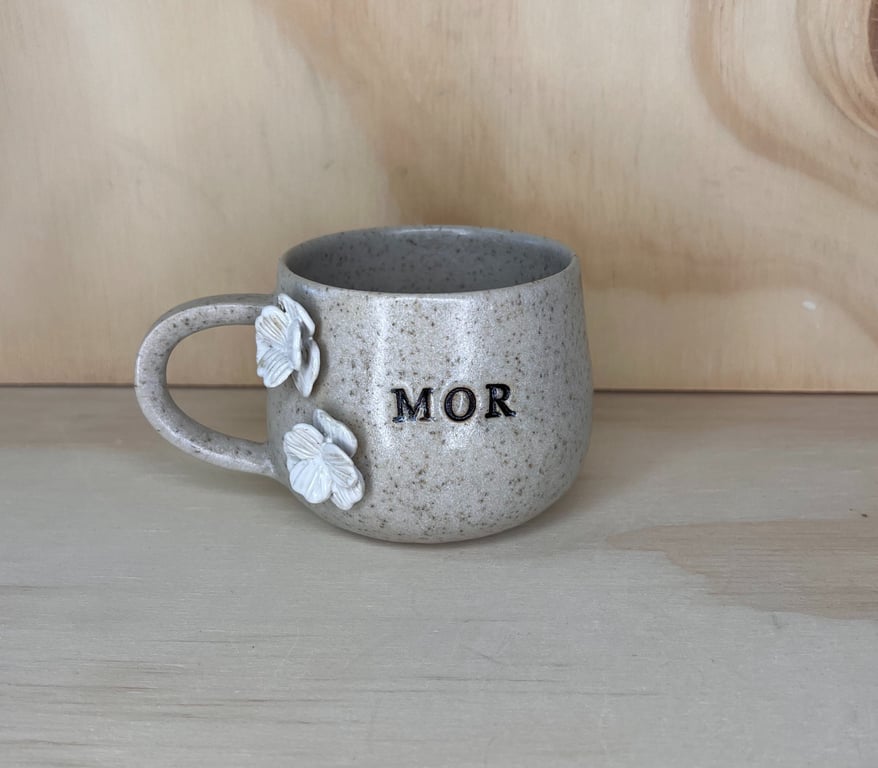 Grå keramikkop med teksten "MOR" og hvide blomstdekorationer på siden, placeret på en lys trækasse.