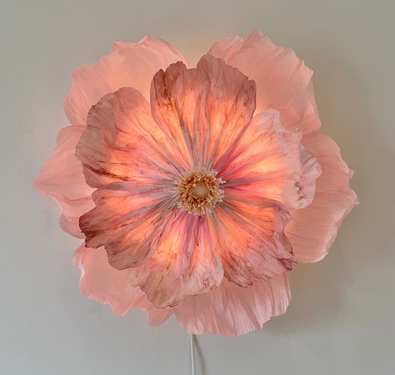 Lysende væglampe formet som en stor lyserød blomst med detaljerede blomsterblade mod en lysegrå væg.