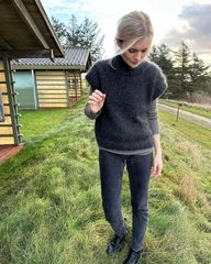Kvinde iført mørke bukser og kortærmet, strikket sweater, står på græs foran hytter under overskyet himmel. (2)