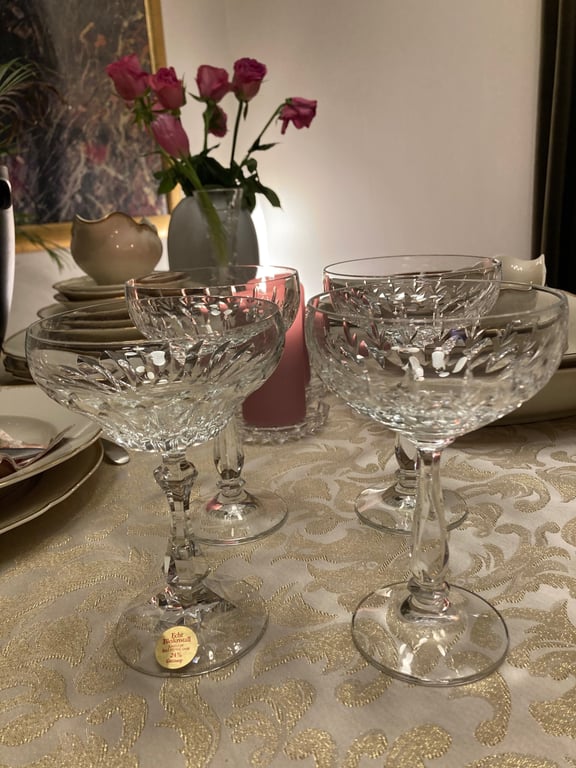 Fire udskårne glas på stilk står på et mønstret dug, med en vase med rosa roser og en lyserød lys i baggrunden. (2)