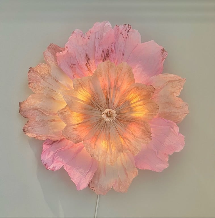 Skærmlys formet som en stor blomst med lyserøde og orange kronblade, der afgiver et varmt, blødt lys mod en hvid væg.