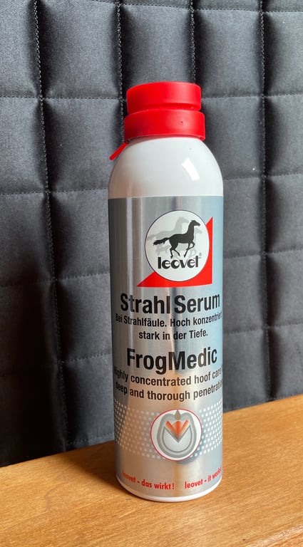 Flaske med Leovet Strahl Serum FrogMedic til pleje af hove med rød låg stående på et træbord foran en grå quiltet baggrund.