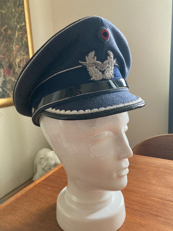 Blå militærkasket med sølvbroderi og sort skygge, placeret på en hvid mannequin hoved på et træbord. (2)