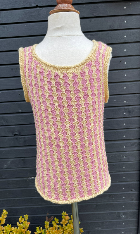 Strikket ærmeløs top i lyserød og beige med mønster, vist på en mannequin foran en sort trævæg. (4)