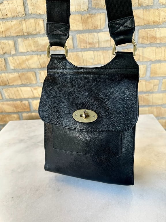 Sort lædertaske med bred sort skulderrem og metallukning foran, placeret på en lysegrå overflade mod en væg af gule mursten.